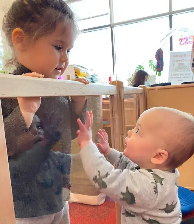 An infant and an older child reach toward each other through a mirrored surface in the Bigelow classroom